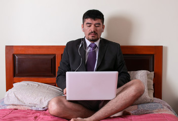 man in formal suit sitting on his bed with headphones on is in a business meeting at his laptop