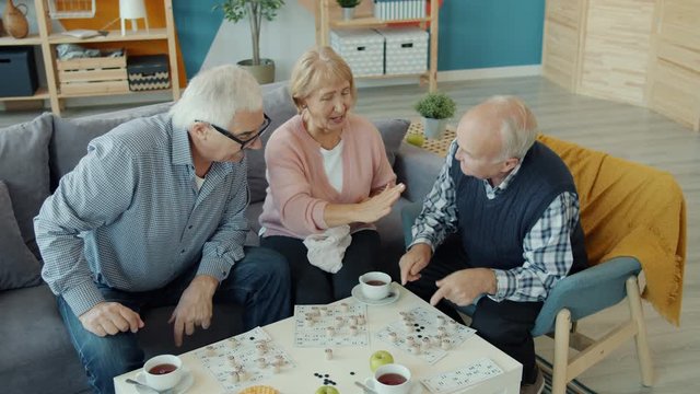 Old men and woman friends are playing lotto doing high-five showing thumbs-up in house. Board games, indoor entertainment and lifestyle concept.