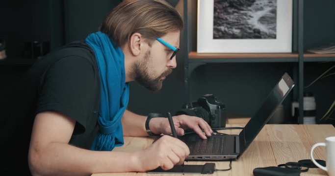 Side view of bearded mature man in stylish outfit retouching images with stylus pen. Serious photographer in eyeglasses using laptop while working in modern studio.