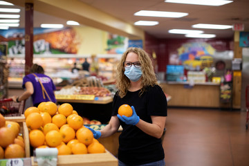A woman who is looking at products like fruit to buy in a grocery store during the pandemic Covid-19 coronavirus pandemic. She is wearing a face mask and latex rubber gloves for protection.
