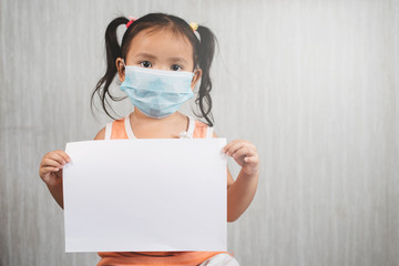 Little asian girl wearing surgical mask while holding blank white paper. Concept of child health care and virus desease outbreak