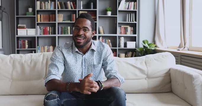 Happy young handsome african american male blogger in glasses sitting on sofa, recording video for followers in social networks. Skilled professional sharing knowledge educating on personal channel.
