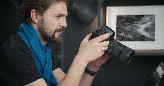 Side view of bearded photographer in blue scarf sitting at table and reviewing images on digital camera. Smiling mature man picking photos for retouching.