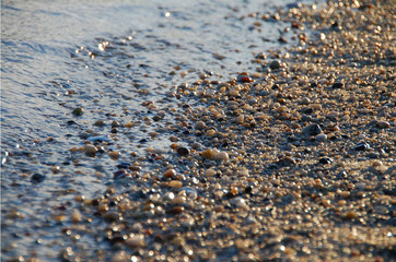 Pebbles on the beach at the ocean at sunset, while the waves lap at the shore on the sandy coast. The sun illuminates the sea and the saltwater on vacation.