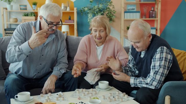 Joyful senior people are enjoying lotto game indoors at home talking entertaining sitting at table with food. Retirement lifestyle and friendship concept.