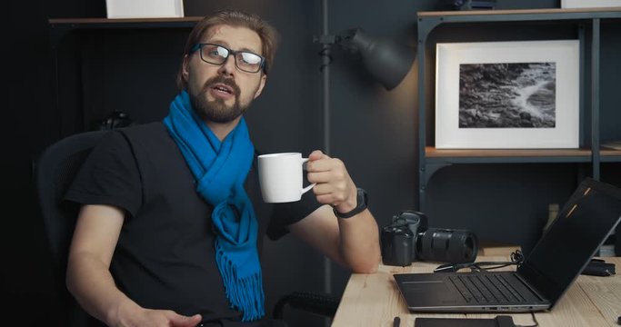 Bearded male instructor sitting at table and drinking coffee while conducting online tutorial about photography. Mature man sharing his knowledge with new photographers.