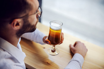 Closeup of young melancholic businessman in shirt sitting in pub next to window and looking trough it. He is holding glass of beer. Selective focus on beer.
