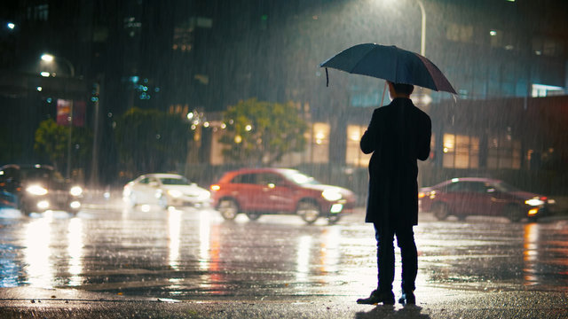 On a rainy night, a man with an umbrella stands on the city street, coronavirus