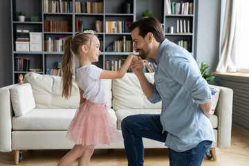 Cute daughter play with father indoor enjoy free time together, dad kneeling holds hand kisses arm...