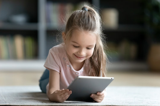Little girl lying on floor in living room with tablet looking at device screen watching cartoons enjoy audiobook listening, preschool modern tech user parental control safety, gadget overuse concept