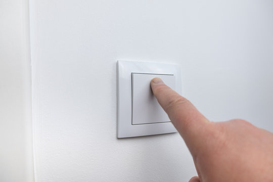 Finger pressing light switch to turn off light. Modern white light switch on white wall.