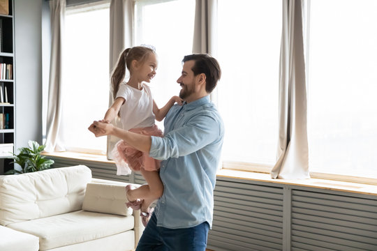 Loving dad holds on hands small daughter she wears crown imagines herself like princess, people dancing in living room, enjoy holiday life event birthday party. Happy fatherhood, family bonds concept
