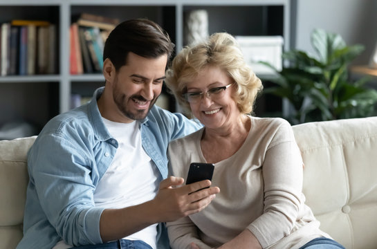 Millennial Adult Son Demonstrates To Older Mother New Application On Smartphone Sit On Couch At Home, Helps Her And Teaches, Shows Old Mom Photos, Share Personal, Having Fun, Modern Tech Usage Concept