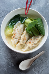 Asian dumpling soup with bok choy served in a white bowl, vertical shot on a grey concrete background