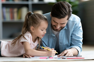 Caring daddy lying on warm wooden floor spend weekend with daughter holding pencils drawing use album close up image, focused little kid girl enjoy time together with loving dad at modern home concept