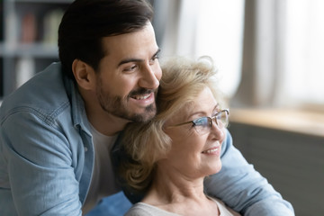 Close up image elderly mother resting sit indoors her grown up son hugs old mom, family looking at distance feels happy spend time together, next generation, care about older relatives people concept
