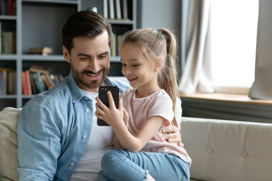 Small daughter seated on father lap showing him cool game on smartphone in internet. Parental control, protect child from forbidden content and access, new generation and mobile electronic fun concept