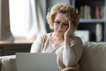 Old woman sit on couch using laptop looks at screen feels stressed not understand need assistance...