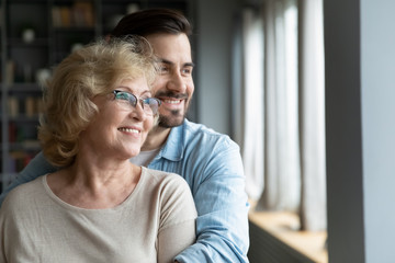 Young adult son hugs elderly mother, family smiling looking out the window standing in living room at home. Offspring and future, grown-up child give care to mom, visit her provide attention concept