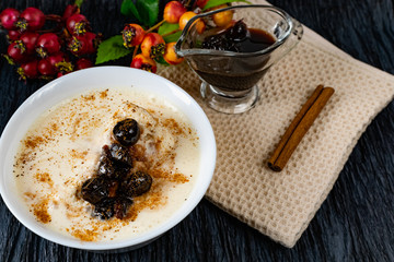 Sour-milk yogurt dessert, with thermostatic cooking, decorated with ripe cherry berries in syrup, on a dark background with jam in sauce and a stick of cinnamon, next to sprigs of fresh red berries
