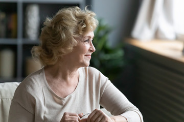 Pretty middle-aged woman sitting on sofa resting at home looking out the window enjoy retired...