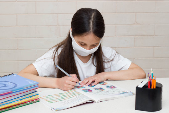 Distance learning online education. A schoolgirl is studying at home and doing homework. Sitting on the bed with textbooks in a protective mask.