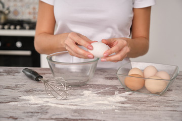 Unrecognizable woman cook in white clothes is going to cook in the kitchen, breaks egg in bowl. On the table is a bowl with eggs, a bottle of milk, a whisk and a flour.