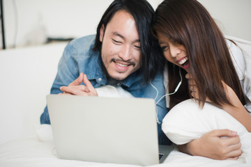 Young Asian man and woman are looking joke movie in laptop computer on the bed at home. the couple man and woman lie on the tummy and smiling with laugh when looking clip vdo. life at home concept.