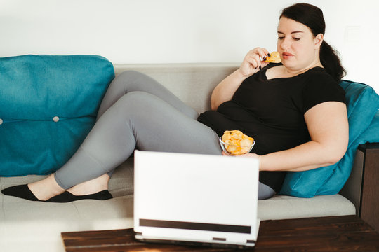 Overeating and gluttony concept. Mindless snacking, home sedentary lifestyle. Obese woman sitting on sofa eating unhealthy food