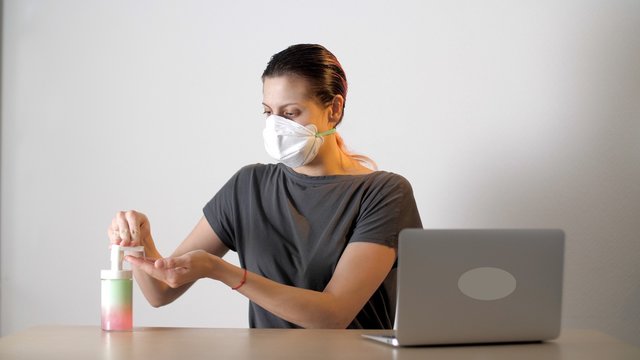 Attractive woman in protective medical mask sitting at the table with laptop. Woman or girl rubs his hands with a sanitizer and works with a laptop. Coronavirus or Covid-19.