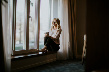 Young woman with cup of tea or coffee sitting and drinking on the window sill at home