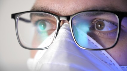 Close-up male face in medical protective mask with glasses. Display reflection in glasses. Portrait of a young man, eyes of programmer with glasses. Work in home. Quarantine