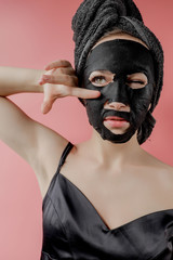 Young woman apply black cosmetic fabric facial mask on pink background. Face peeling mask with charcoal, spa beauty treatment, skincare, cosmetology. Close up