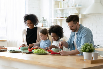 Happy young multiethnic family with little daughters have fun cooking at home together, smiling parents and small multiracial children prepare breakfast salad in kitchen, healthy lifestyle concept