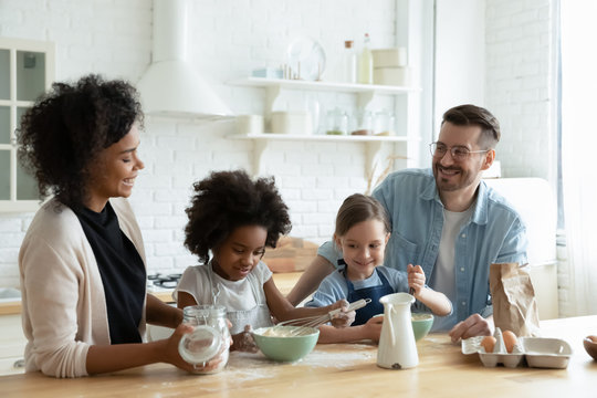 Overjoyed diverse parents have fun bake in modern kitchen with little multiethnic daughters, happy multiracial young family with small girls children prepare biscuits pancakes for breakfast at home