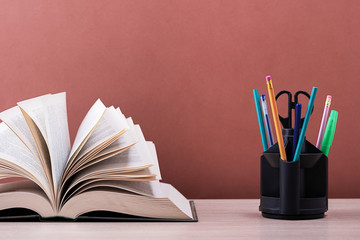 A large thick book with the pages spread out like a fan and a stand with pens, pencils and scissors on the table on a brown background.