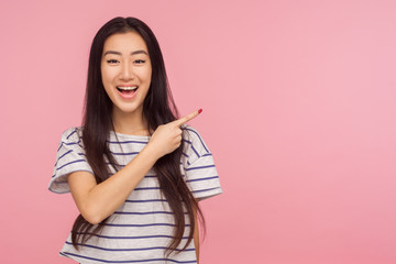 Look at advertisement! Portrait of charming asian girl in striped t-shirt pointing to side and smiling, showing copy space, empty place for commercial. indoor studio shoot isolated on pink background