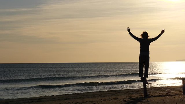 Silhouette of woman at dawn morning on beach, looking at sea horizon, enjoying sunrise with arms raised up. Back view