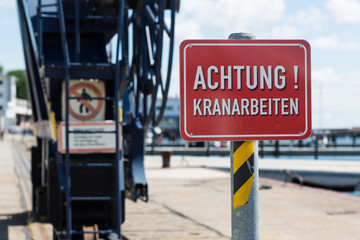 a red sign "Attention! Crane work" (German "Achtung! Kranarbeiten") in front of crane constructions on a quayside