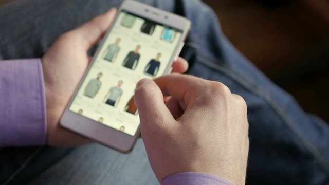 Man Looks at Goods in Online Store Clothing. Buy Fashion Clothes Directly on Smartphone. Man at Home in Living Room Using Smartphone. Screen is blurred. Focus on hand.