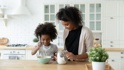 Obraz na płótnie Canvas Loving young African American mother teach small biracial daughter bake in kitchen, happy caring ethnic mom and little girl child preparing pancakes or biscuits, make breakfast at home together