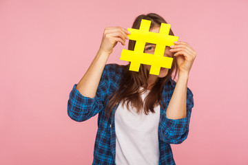 Happy playful girl in checkered shirt hiding behind yellow hashtag symbol, covering face with hash...