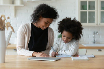 African American young mother and little daughter sit at desk in kitchen studying online together,...