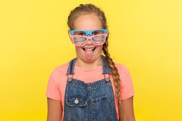 Fashionable eyeglasses. Portrait of little girl wearing two stylish eyeglasses and showing tongue making comical face, child having fun with accessory. indoor studio shot isolated on yellow background