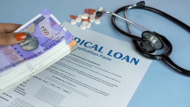 Closeup shot of a blank Medical loan application form - personal loan concept. Pan shot of woman hands placing a bundle of Indian currency notes next to colorful pills and a stethoscope - blue back...