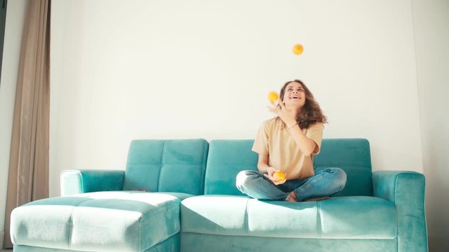 A young beautiful woman juggling with three oranges while sitting on the couch in the living room. Self-isolation and lockdown concept.