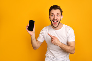 Funny young man in white t-shirt on yellow background, presenting smart phone