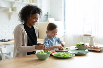 Happy young biracial mother and little Caucasian daughter chop vegetables prepare healthy salad at home kitchen, smiling African American mom and small girl child cooking making breakfast together