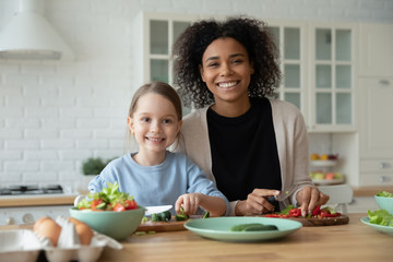 Portrait of smiling African American mom and little Caucasian daughter look at camera cooking in kitchen together, happy biracial mother and small girl child have fun preparing healthy food at home