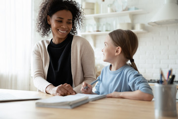 Smiling young biracial mom help little daughter with homework in home kitchen, happy caring mother study together with small girl child handwriting, having online lesson, homeschooling concept